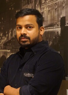 Herr Kumar Kizhake Malaily mit verschränkten Armen, vor urbanem Schwarz-Weiß-Hintergrund.